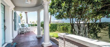 A covered patio with white columns, a brick floor, and a view of a lush garden and a body of water.