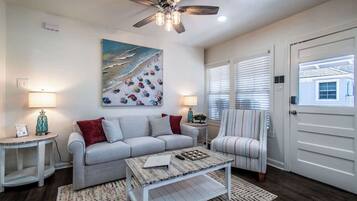 Un salon avec un canapé gris, une table basse, un ventilateur de plafond et un tableau représentant une scène de plage.