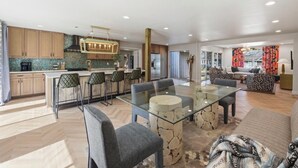 A modern kitchen with a central island, dining area with a glass table, and a living room with a sofa and patterned cushions.