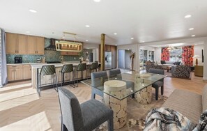 Una cocina moderna con una isla central, comedor con mesa de vidrio y sala de estar con sofá y cojines estampados.