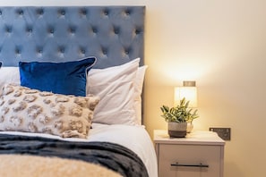 A neatly made bed with a tufted headboard, a bedside lamp, and a potted plant on the nightstand. - Creative Quarter Luxury 2 Bed City Apt (Birmingham)