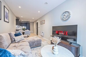 A modern living room with a sofa, a coffee table, a wall-mounted clock, and a large TV screen displaying the Netflix logo. - Creative Quarter Luxury 2 Bed City Apt (Birmingham)