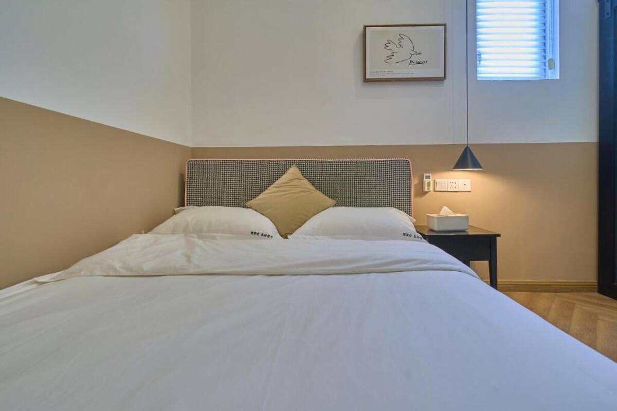 Een net opgemaakt bed met witte lakens en een hoofdbord, een nachtkastje met een lamp, en een ingelijst beeld aan de muur.