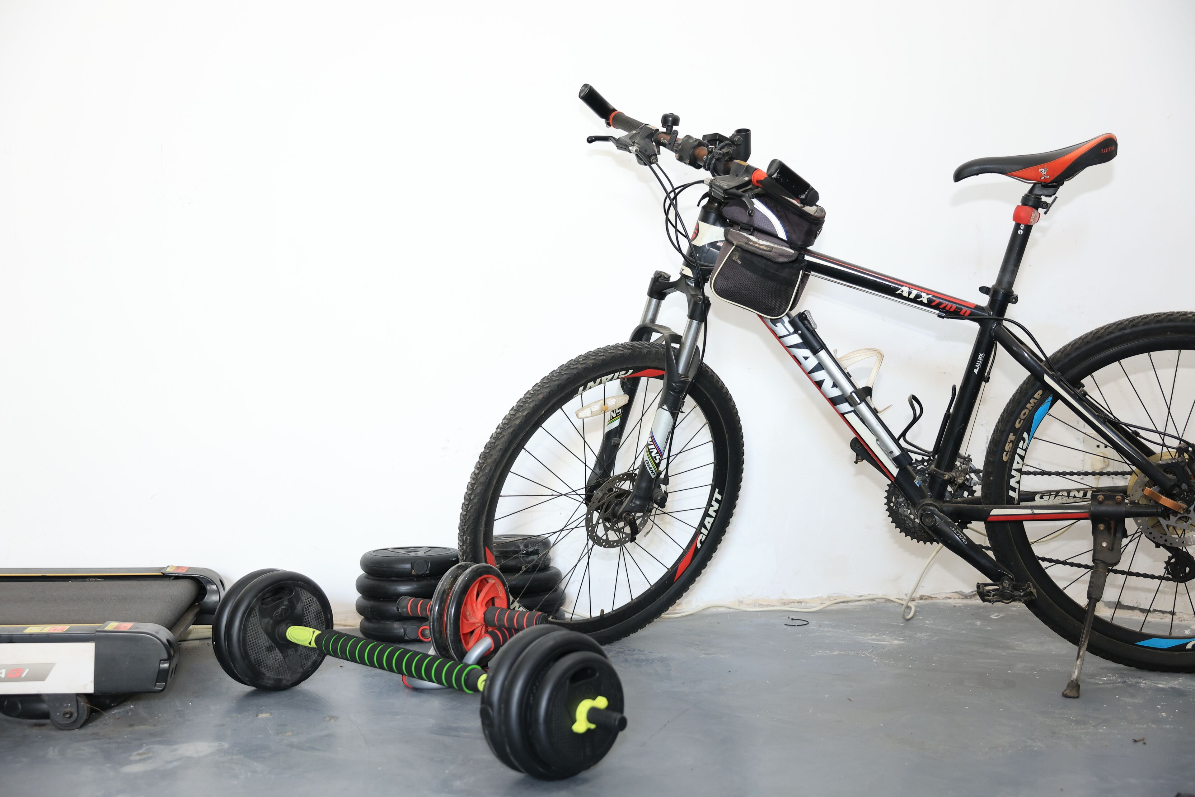 Una bicicleta con estructura negra y detalles rojos, un juego de mancuernas y una cinta de correr en una habitación con una pared blanca lisa.