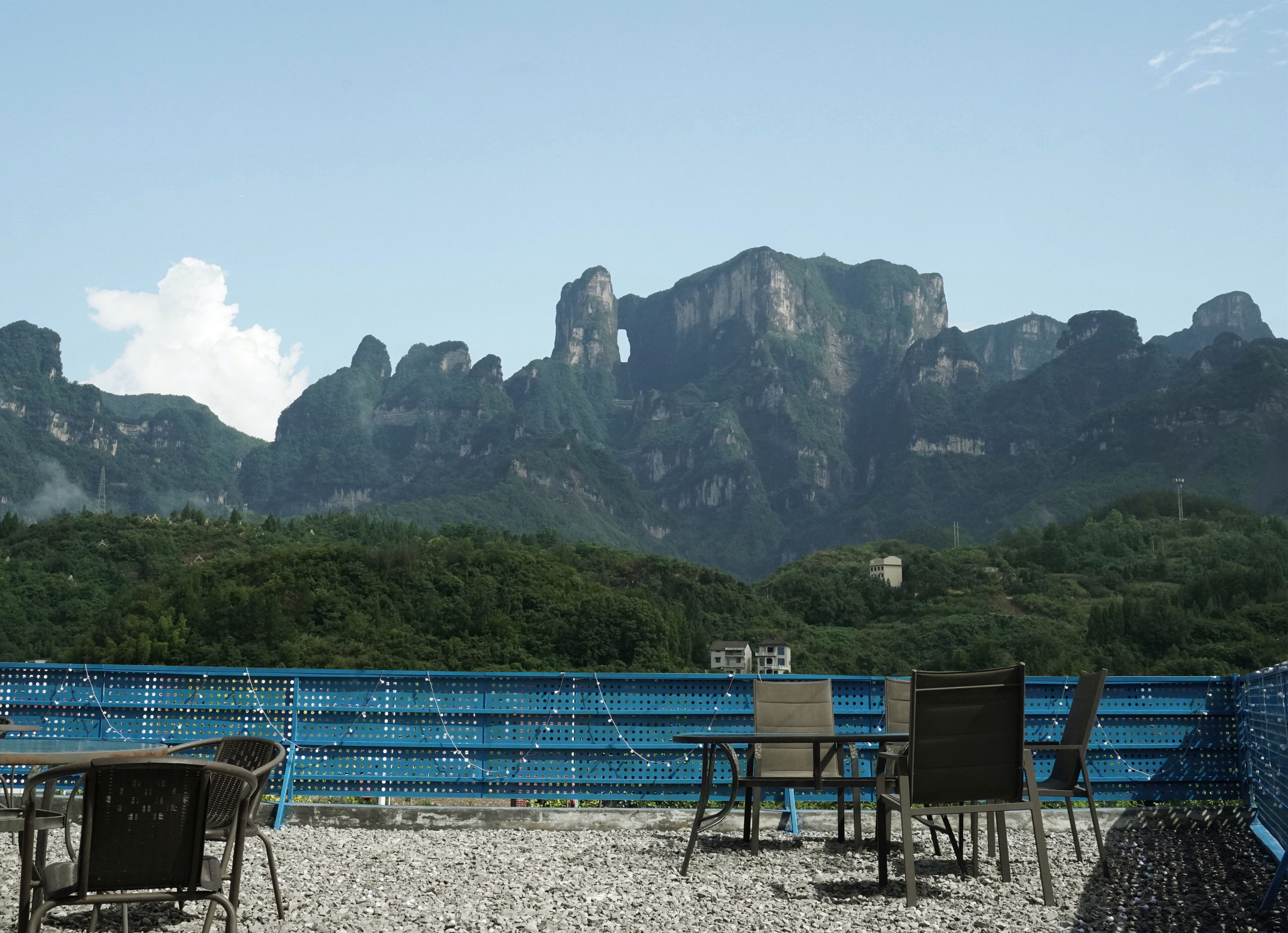 Una terraza con sillas y una valla azul, con vistas a un paisaje montañoso donde se divisa un edificio a lo lejos.