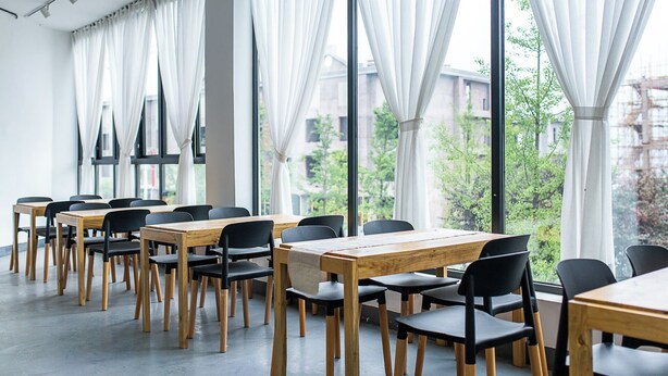 A conference room with tables and chairs, large windows with curtains, and a view of buildings and trees outside.