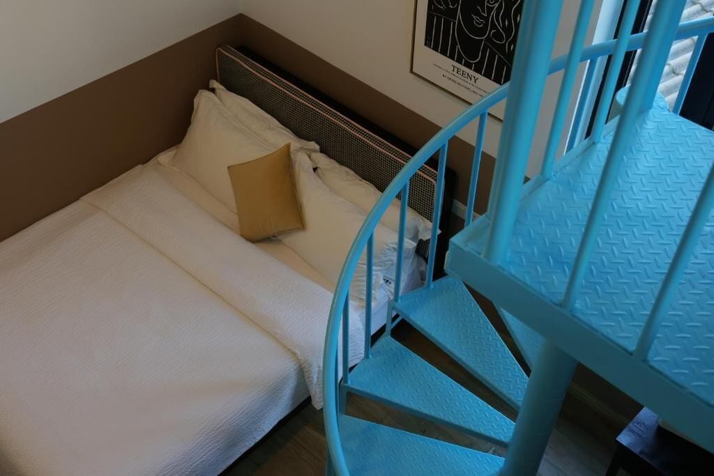 Een bed met wit beddengoed en een bruin kussen, een blauwe metalen stoel en een lijst met een afbeelding aan de muur.