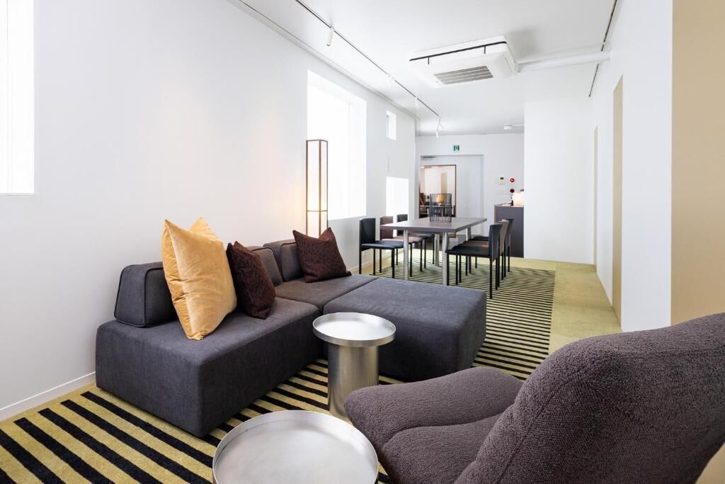 Ett modernt hotellrum med en hörnsoffa, ett runt sidobord och en matplats i bakgrunden.