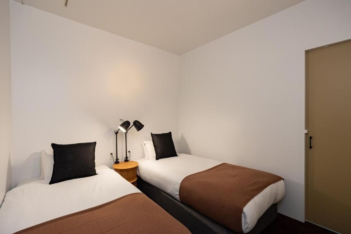 A hotel room with two beds, a bedside table with lamps, and a door.