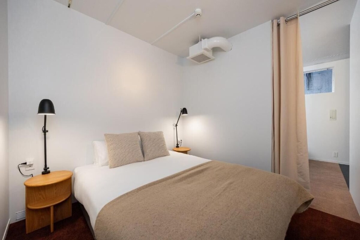 A bedroom with a bed, two bedside tables with lamps, a window with curtains, and a white ceiling vent.