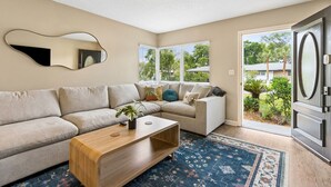 A living room with a sectional sofa, a coffee table, a mirror, and a view of the outdoors. - Blake's Place: Quiet 3bd/2ba home with Mega Hot Tub + Cold Plunge (Jacksonville Beach)