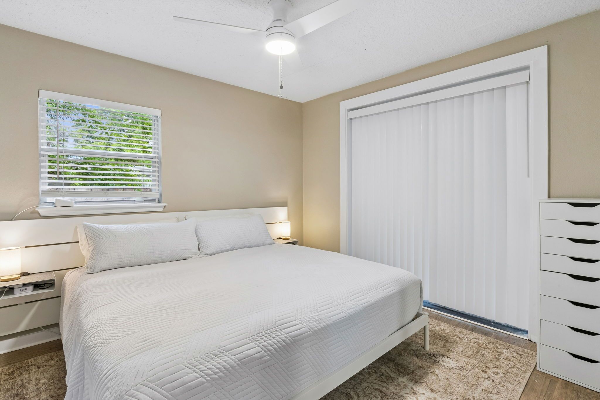 Un dormitorio con cama, mesitas de noche, ventilador de techo, ventana con persianas y armario blanco.
