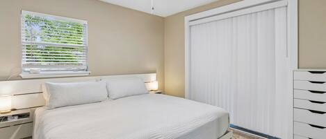 Un dormitorio con cama, mesitas de noche, ventilador de techo, ventana con persianas y armario blanco.