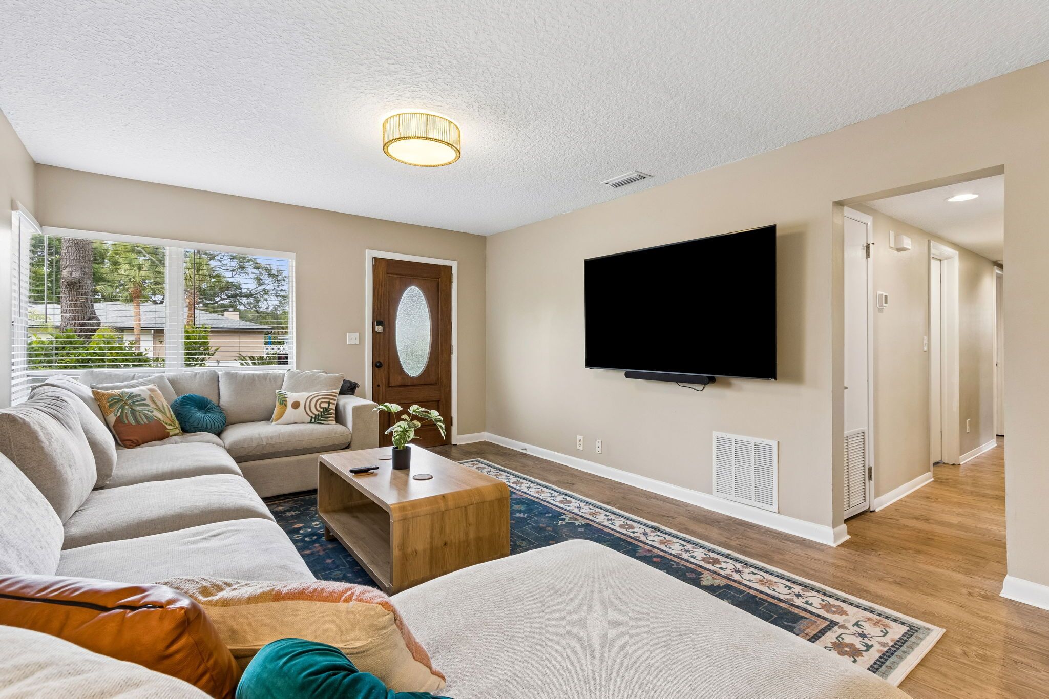Una sala de estar con un sofá modular, una mesa de centro, un televisor de pantalla plana montado en la pared y un amplio ventanal con vistas a árboles y una casa.