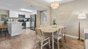 A modern kitchen with white cabinets, stainless steel appliances, a dining area with a wooden table and wicker chairs, and a floor lamp. - Blake's Place: Quiet 3bd/2ba home with Mega Hot Tub + Cold Plunge (Jacksonville Beach)
