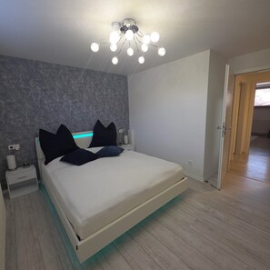 Quarto com cama, mesas de cabeceira, luminária no teto e uma porta.