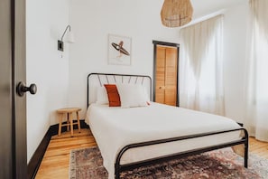 Een slaapkamer met een bed, een klein krukje, een wandlamp, een deur en een raam met gordijnen.