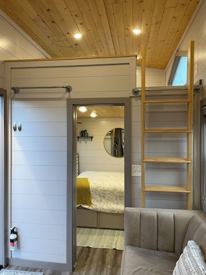 Ein Zimmer mit einem Bett, einer Holzleiter, einem Spiegel und einer Tür zu einem weiteren Raum.
