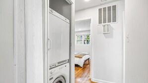 A laundry room with a washing machine, a bed with a white cover, and a window with a view of greenery.