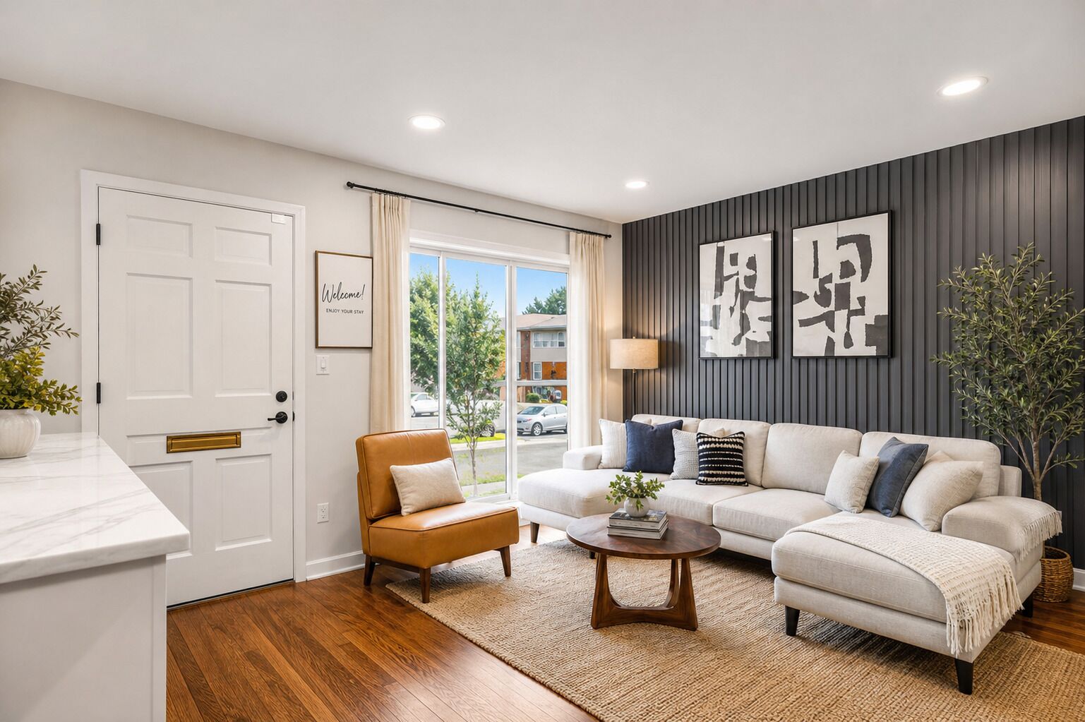 Uma sala de estar moderna com sofá, poltrona e mesa de centro, uma porta branca com uma placa de 'Welcome' e vista para a rua através de uma janela grande.
