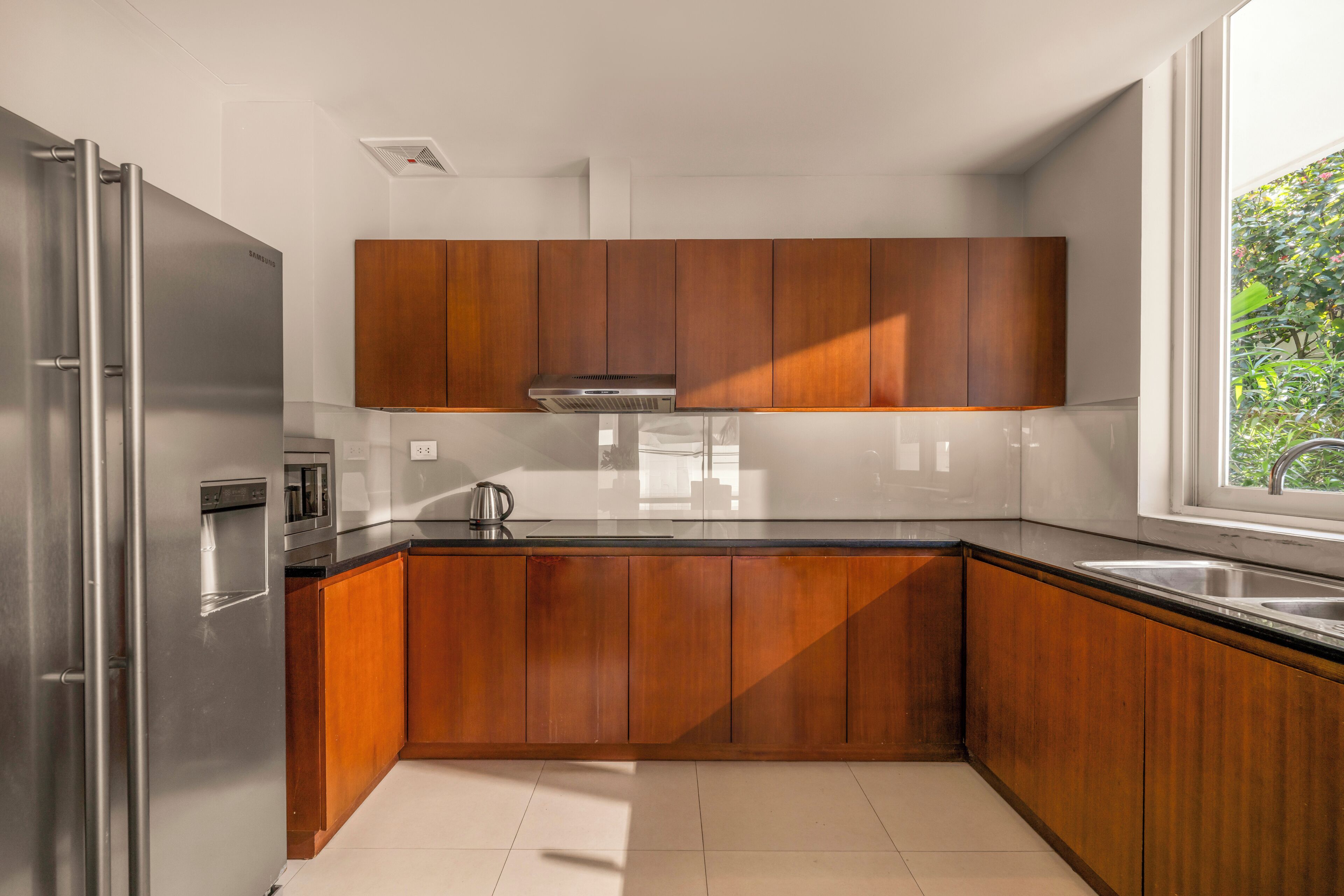 Cocina moderna con armarios de madera, electrodomésticos de acero inoxidable y ventana con vistas a la vegetación.