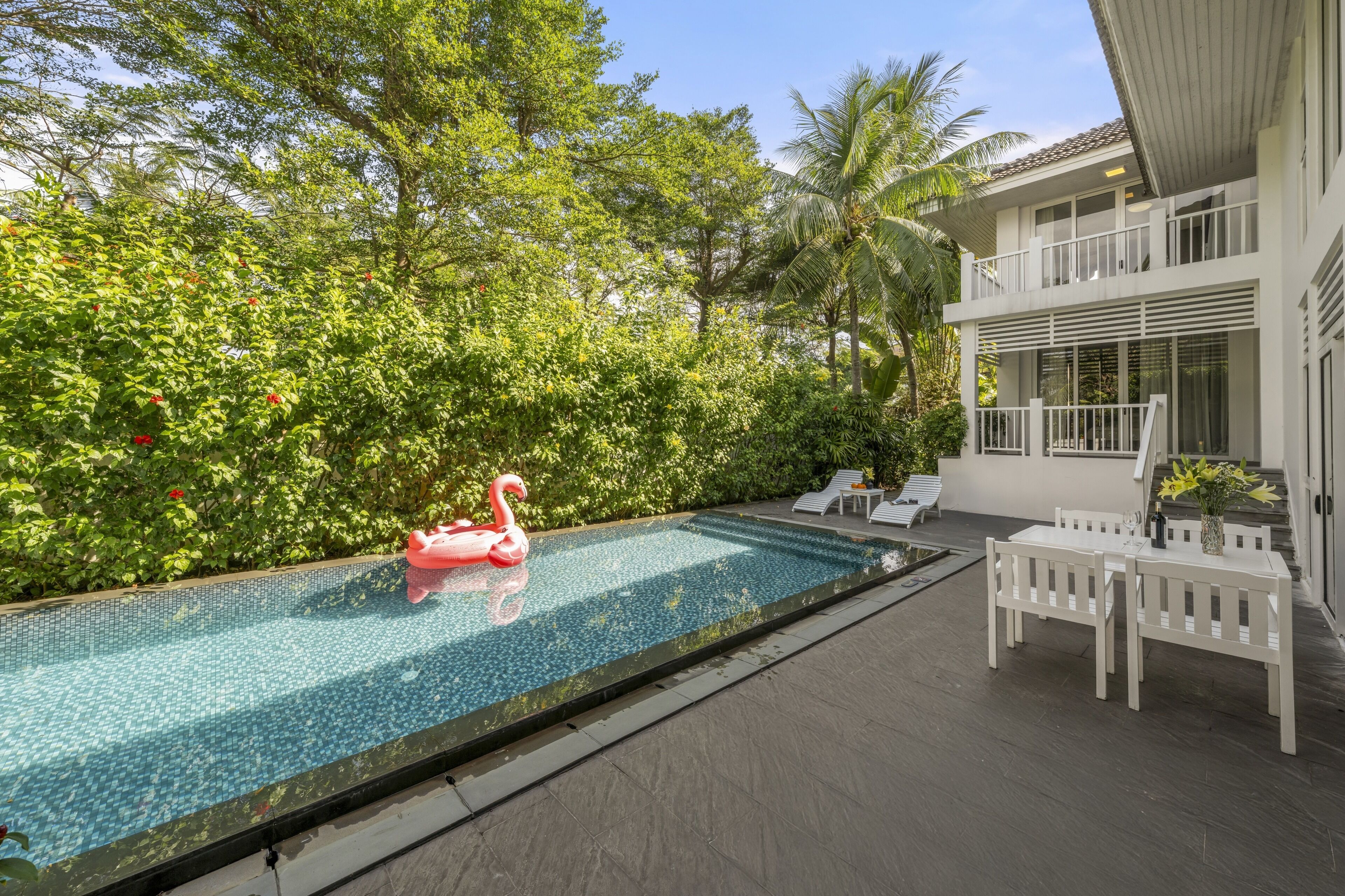 A swimming pool with a pink flamingo float, surrounded by lush greenery and a modern building with a balcony.