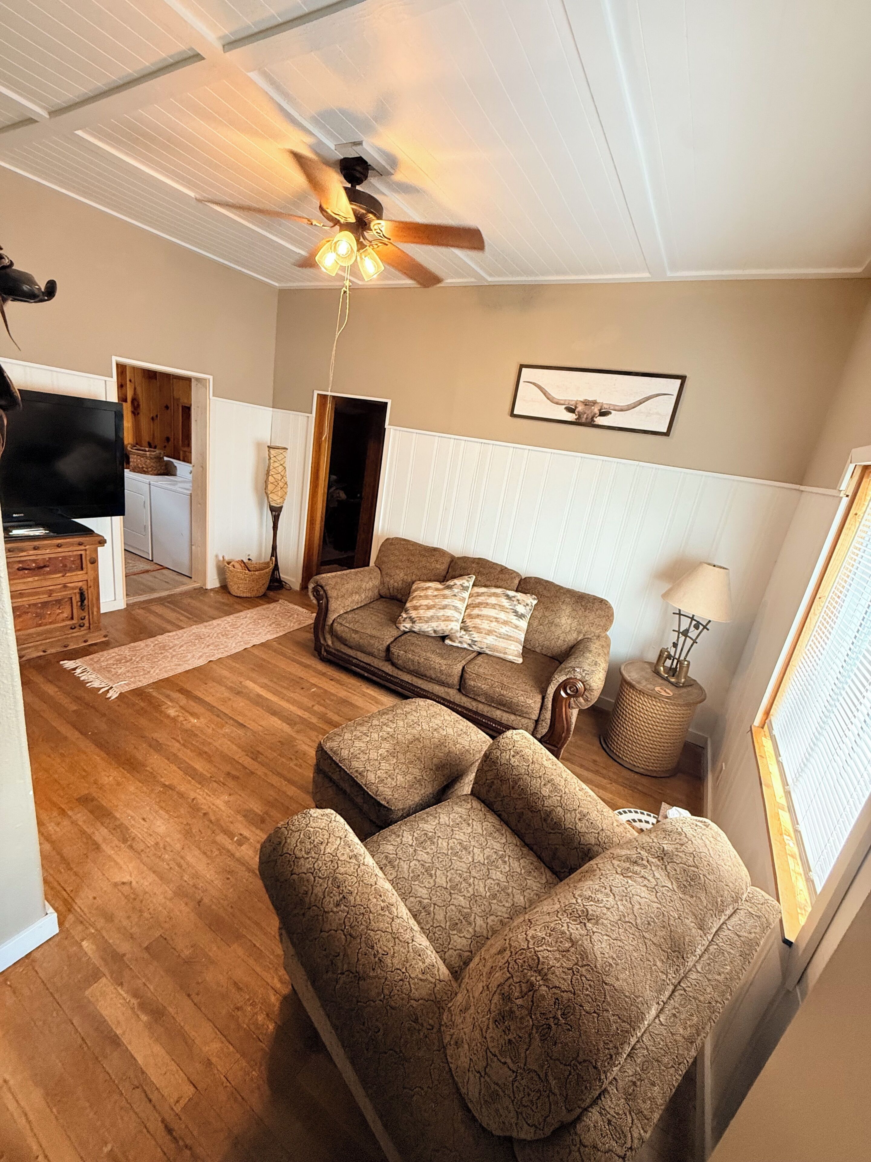 A living room with a sofa, ottoman, ceiling fan, and a television.