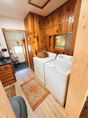 A compact kitchen with wooden walls, a washing machine, a dryer, a small window, and a rug.