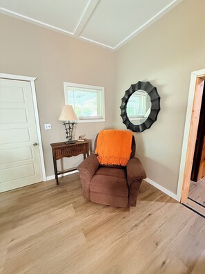 A room with a brown armchair, a small wooden table with a lamp, a window with blinds, and a decorative mirror on the wall. - Cozy  2-bedroom house in beautiful Sundance Wyoming perfect for relaxing stays (Sundance)