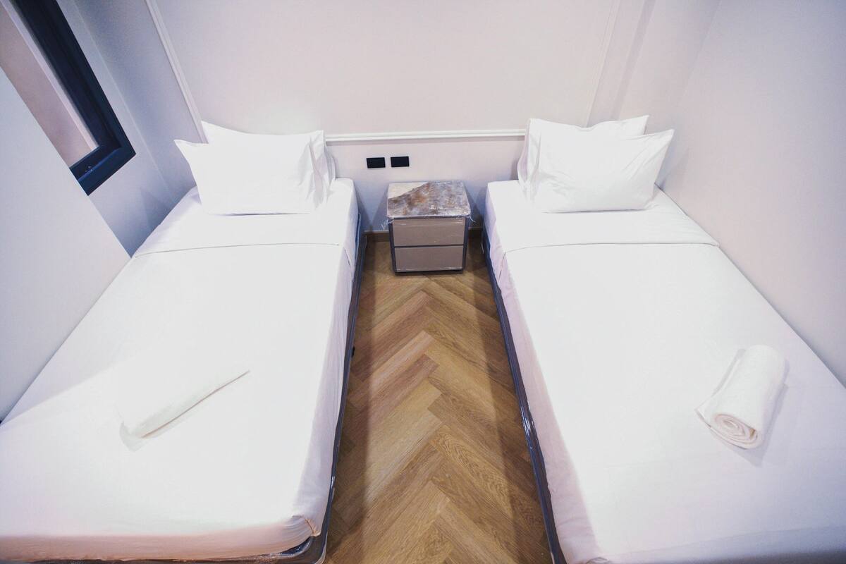 A small, clean room with two single beds, a wooden floor, a small bedside table, and a window.