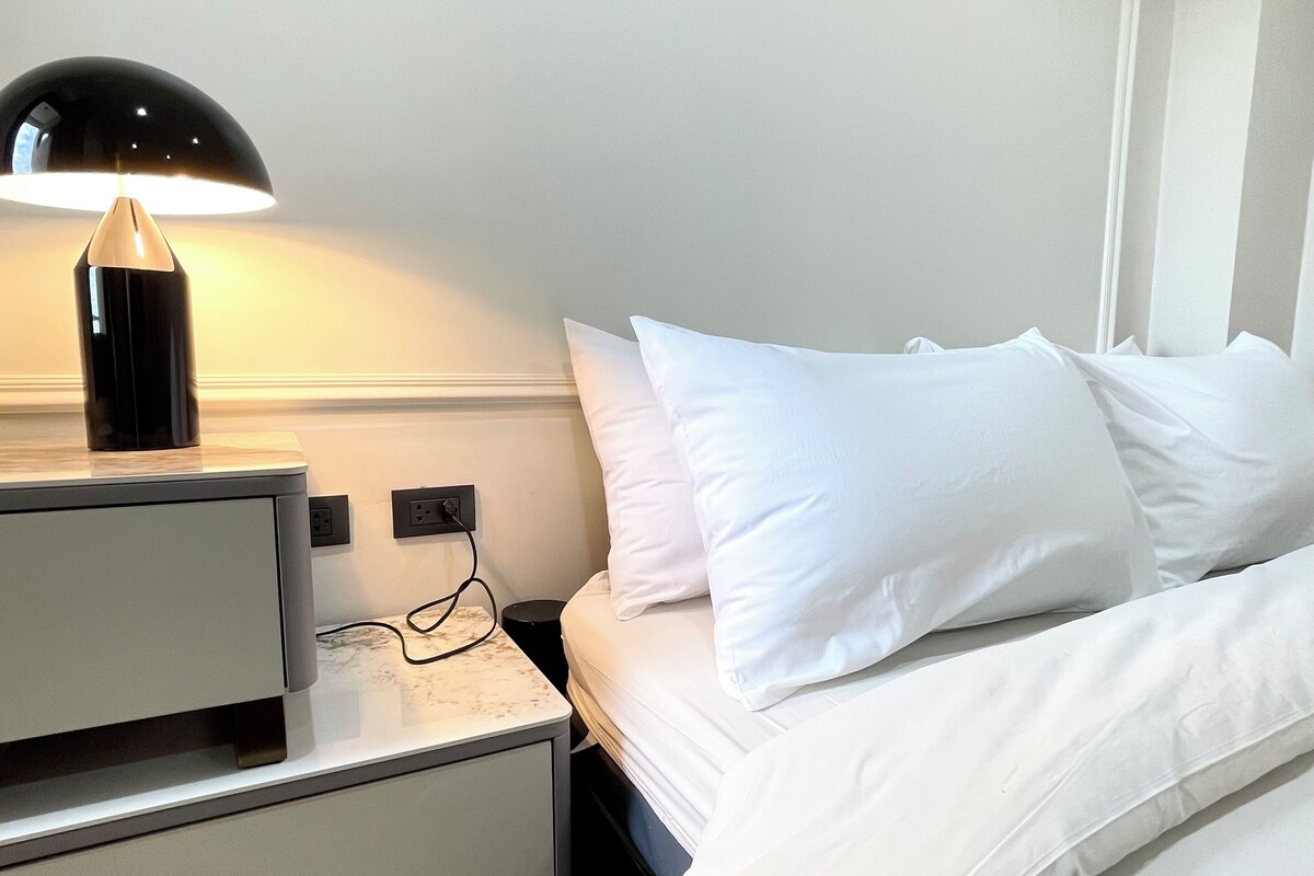 Habitación de hotel con una cama, ropa de cama blanca, una mesita de noche con lámpara y una cómoda.