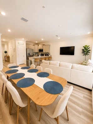 Eine moderne Küche mit Kochinsel, ein Essbereich mit Holztisch und weißen Stühlen sowie ein Wohnzimmer mit weißem Sofa und Flachbildfernseher.