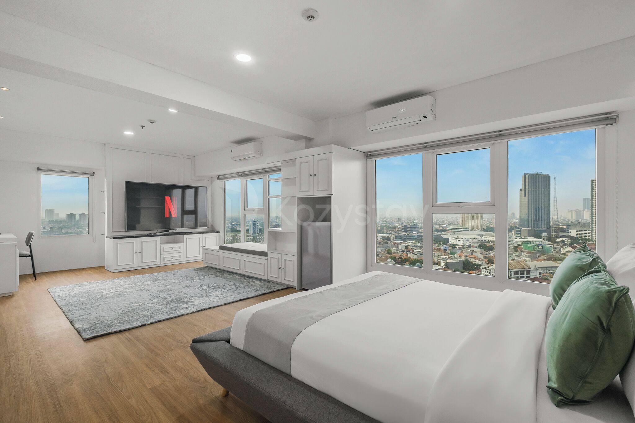 A modern bedroom with a large bed, a view of the city, a kitchen area, and a seating area.