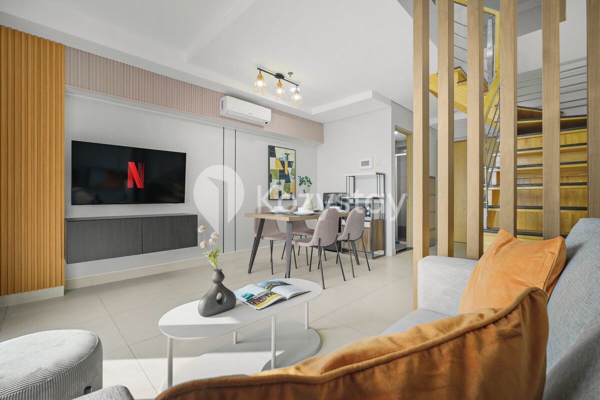 A modern living room with a flat-screen TV, a dining area with chairs and a table, and a staircase with wooden railings.