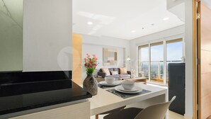 A modern kitchen with a black countertop, a dining area with a table and chairs, a sofa, and a large window with a city view. - Magnolia by Kozystay - Kebon Jeruk (Jakarta Selatan)