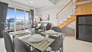 A modern dining area with a glass table, grey chairs, and a city view through large windows. - Magnolia by Kozystay - Kebon Jeruk (Jakarta Selatan)