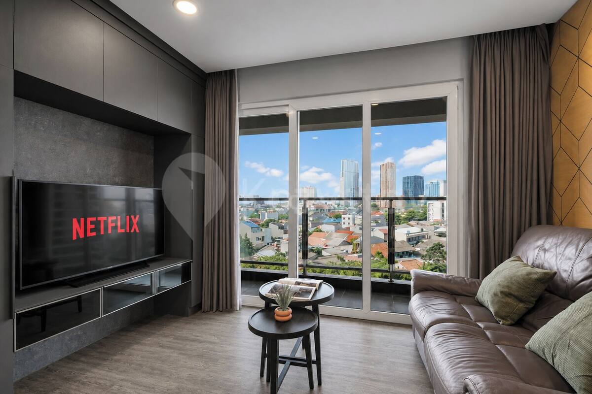 Una sala de estar moderna con un televisor de pantalla plana grande, un sofá, una mesita con una planta y vistas a un paisaje urbano a través de ventanas amplias.