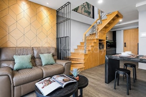 A modern living room with a leather sofa, a coffee table with magazines, a wooden staircase, and a kitchen area.