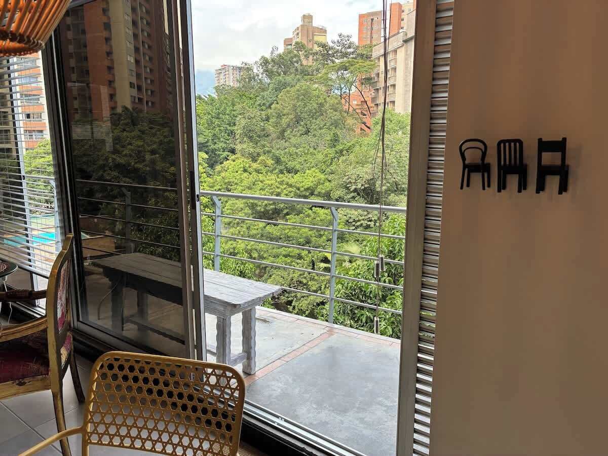 A balcony with a table and chairs, a view of greenery and buildings, and a wall with a chair symbol.