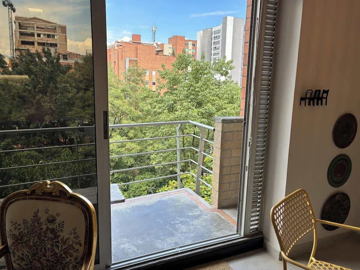 A balcony with a view of buildings and trees, a chair, and a decorative plate on the wall.
