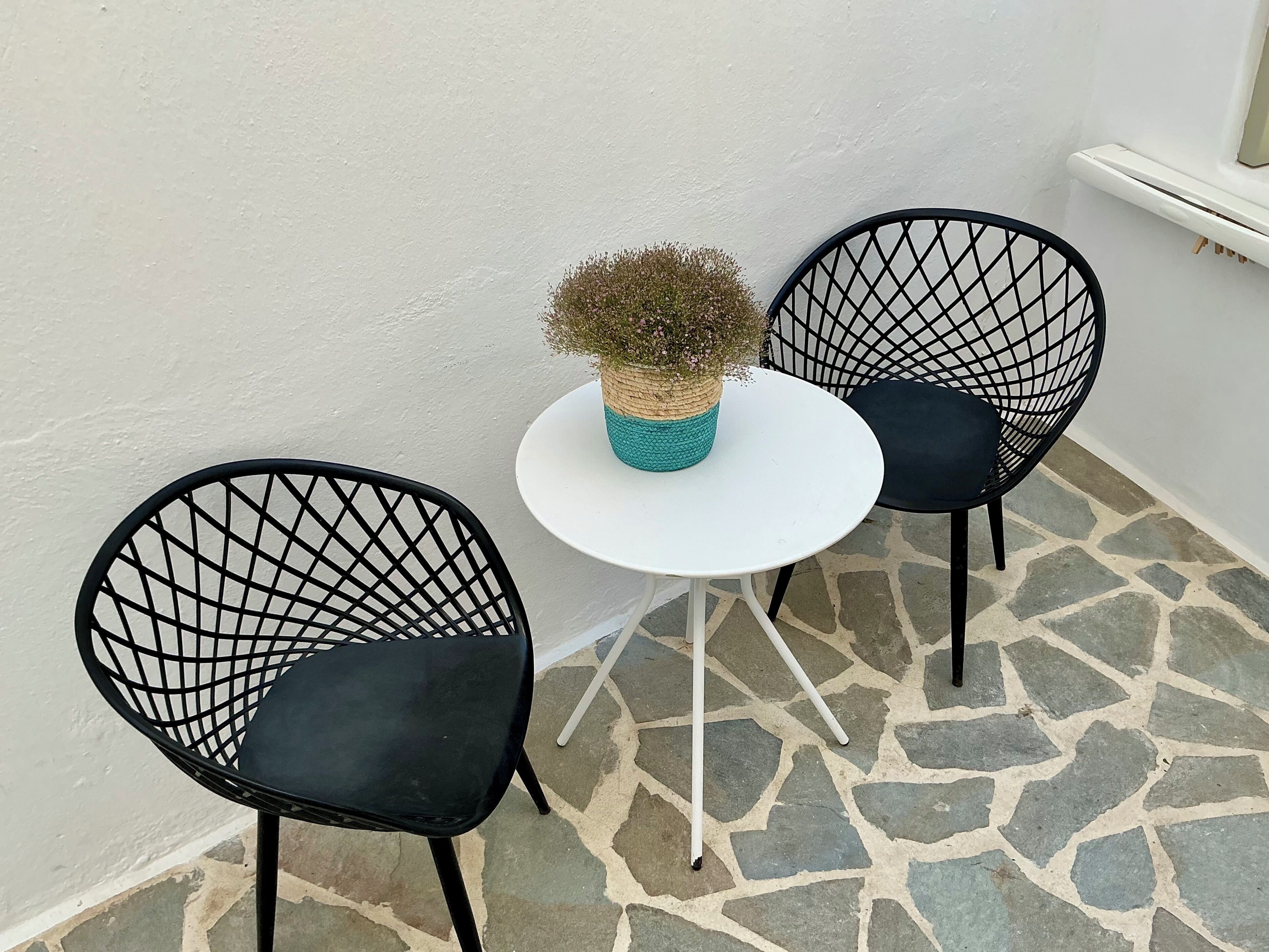 Un petit espace extérieur aménagé avec deux chaises en fil de fer noir et une table blanche, agrémenté d’un pot de fleurs en fibres tressées.