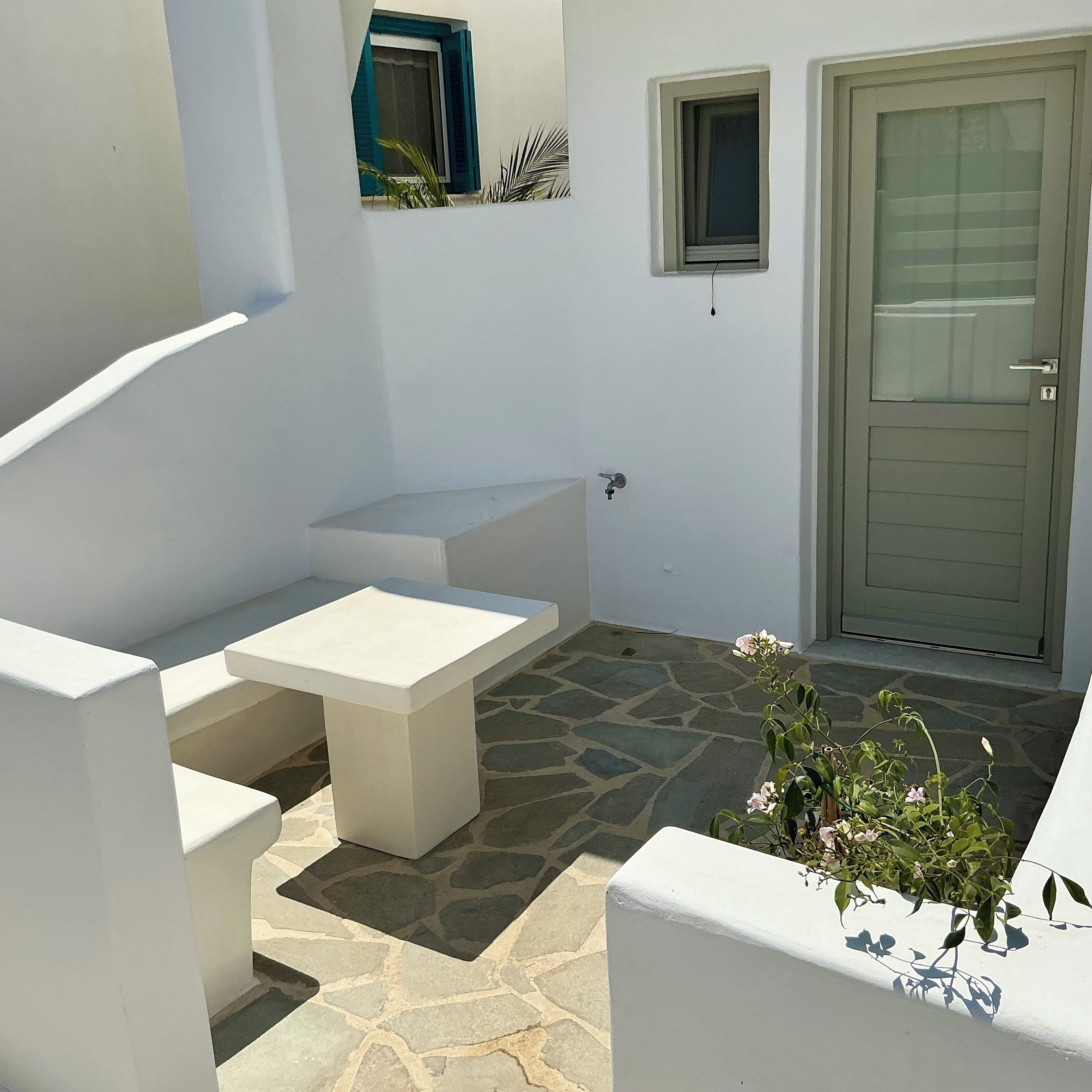 A white outdoor seating area with a stone floor, a door, and a small window with blue shutters.