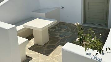 A white outdoor seating area with a stone floor, a door, and a small window with blue shutters.