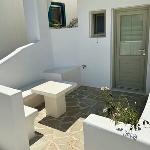 A white outdoor seating area with a stone floor, a door, and a small window with blue shutters. - Ione Inn (Chora)