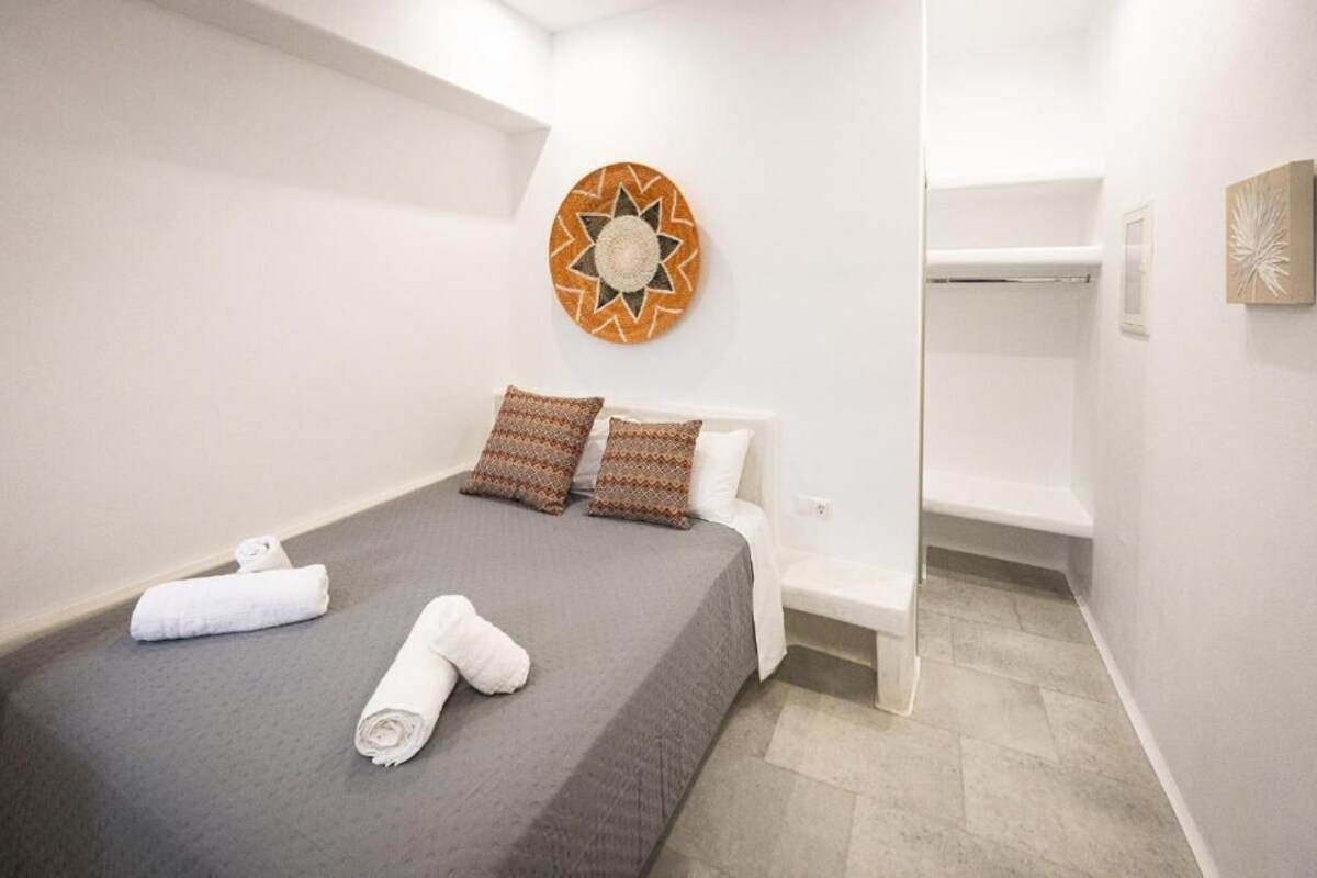 Cama bem arrumada com colcha cinza e duas almofadas estampadas, uma toalha branca e um objeto decorativo na parede.