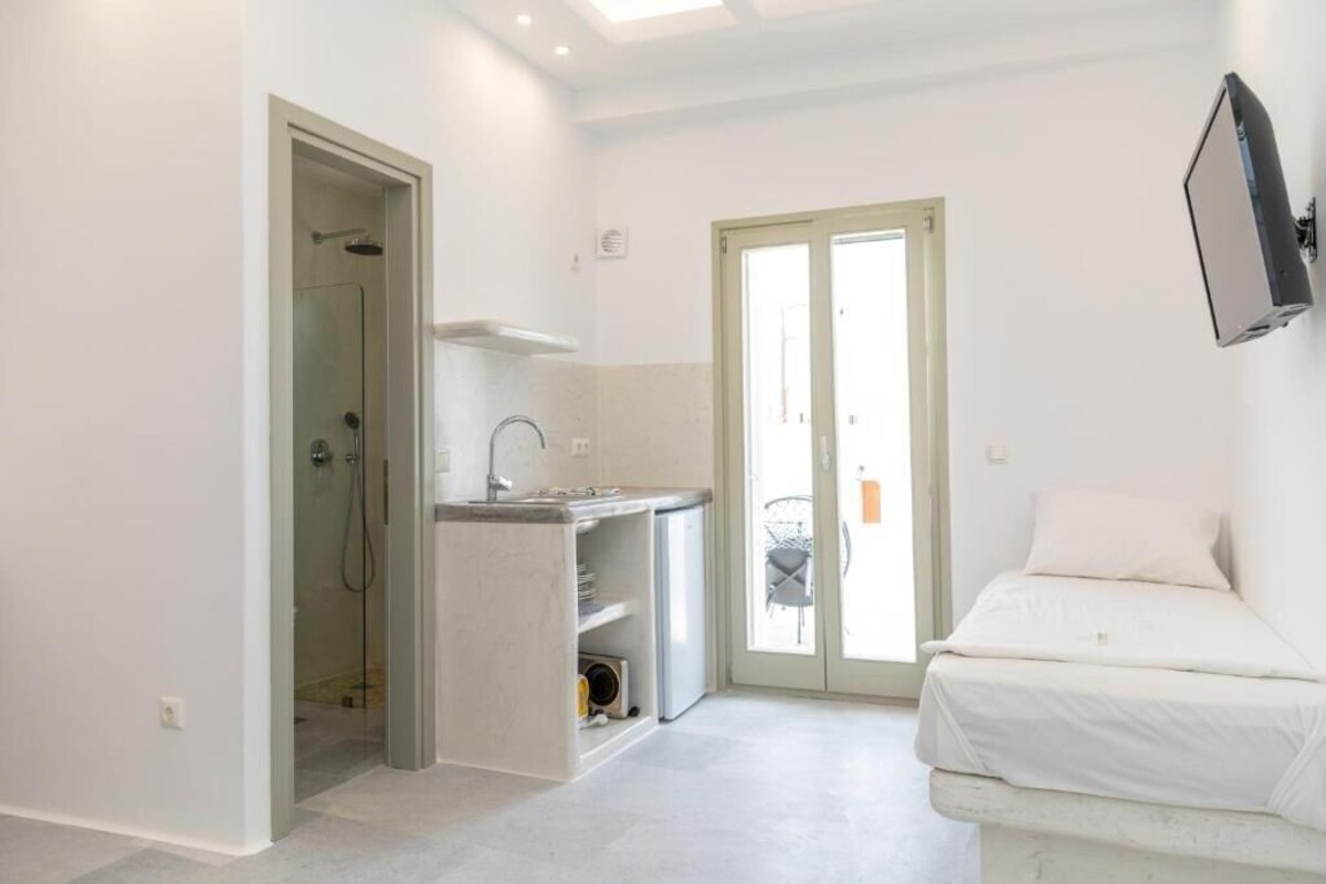 Quarto compacto com cama, cozinha compacta, chuveiro e porta de vidro deslizante para uma varanda.