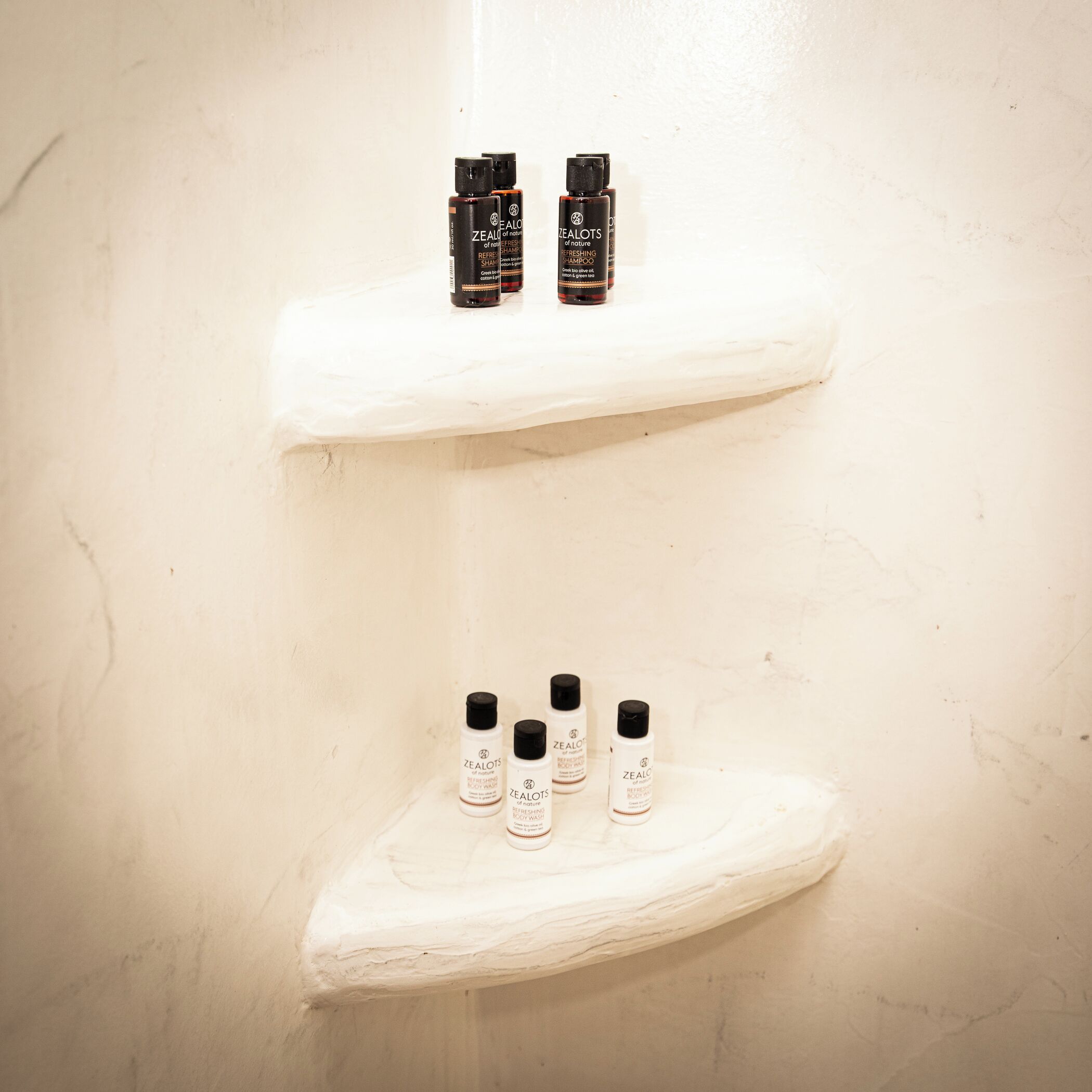 Two white shelves against a wall, each holding bottles of Zealots body wash and shampoo.