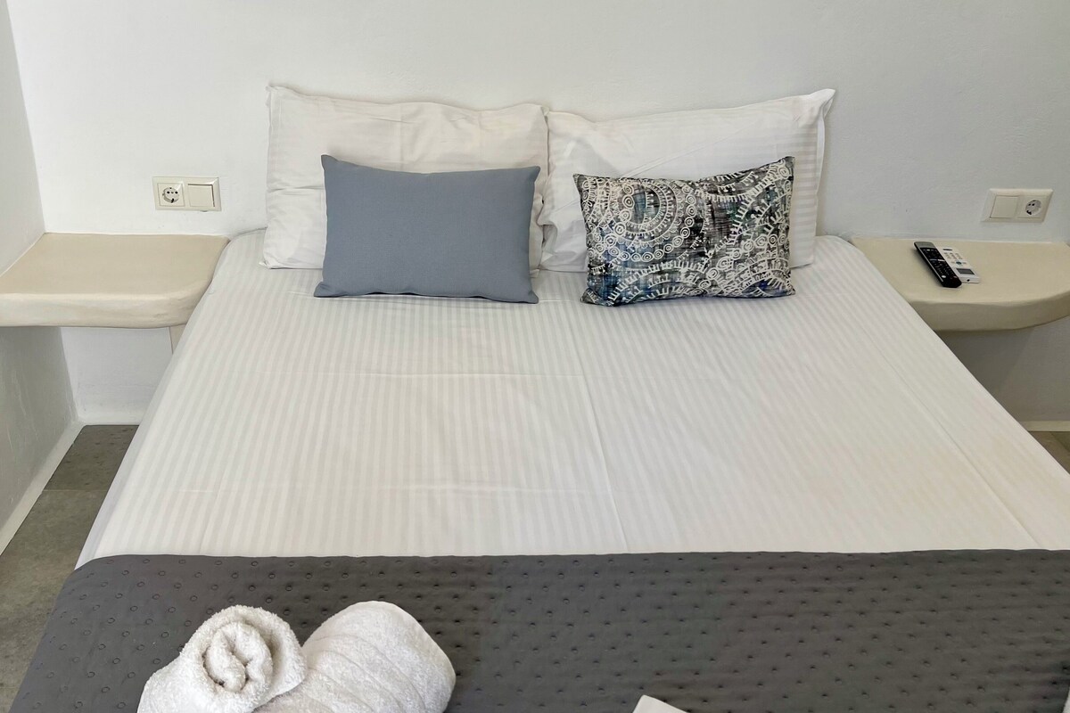Quarto de hotel com uma cama, duas almofadas, mesa de cabeceira com um controle remoto e uma toalha enrolada no chão.