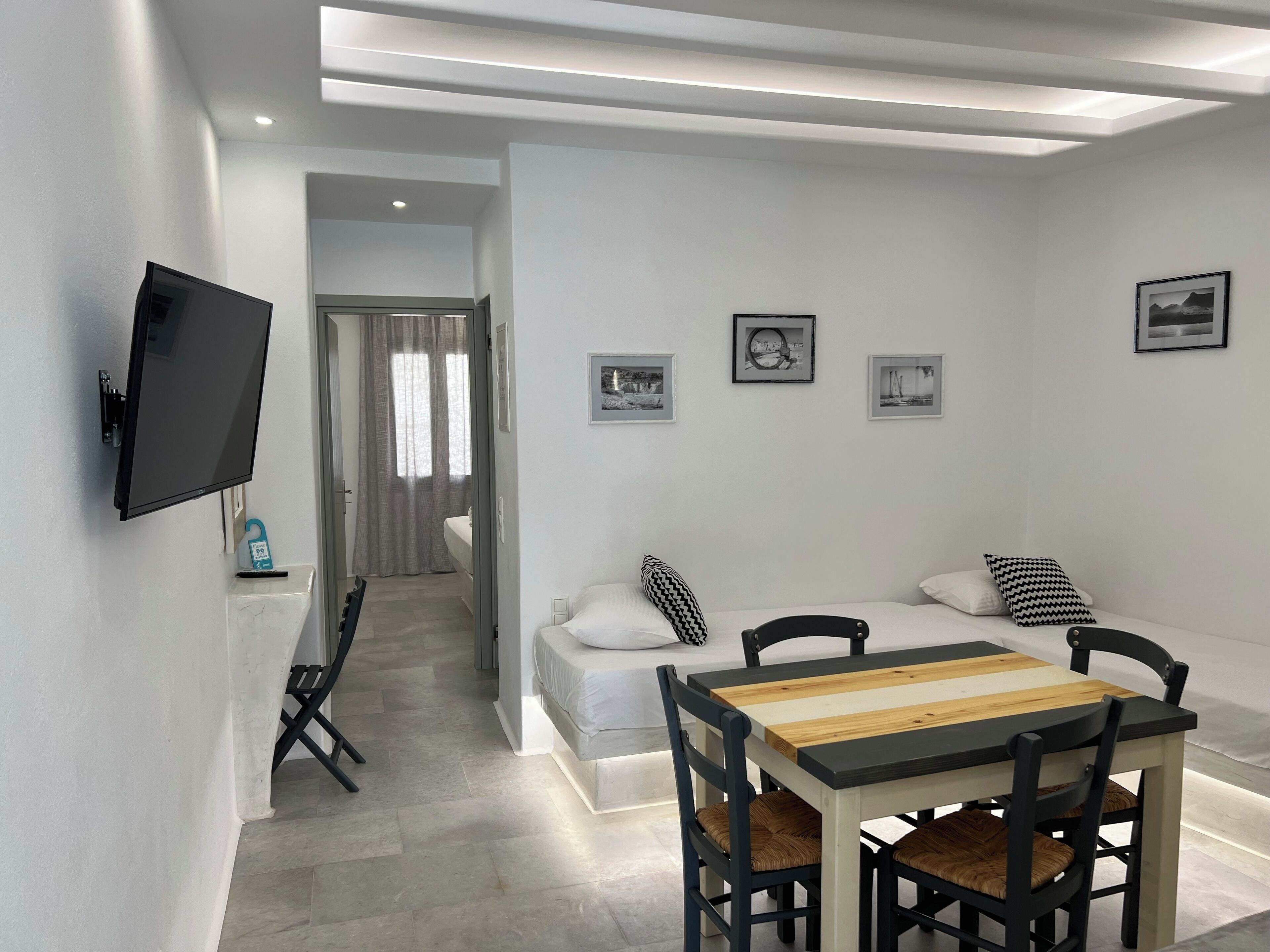 A modern, minimalist room with a flat-screen TV, a dining table with chairs, a sofa with cushions, and framed pictures on the wall.
