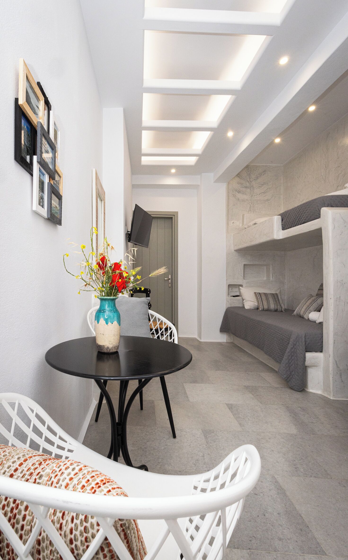 Habitación pequeña con literas, una mesa redonda de comedor con sillas, un florero con flores y un televisor de pantalla plana montado en la pared.